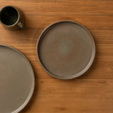 Two round ceramic plates in a matte taupe-brown glaze are arranged on a warm wood tabletop, with a matching dark ceramic mug partially visible in the upper left.