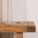 A round travertine stone tray with a natural stone edge, displayed on a rustic wooden table next to a beige linen curtain.