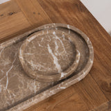Two round marble trays in a warm taupe-brown color with white veining, nested inside a larger matching rectangular tray, displayed on a wooden table.