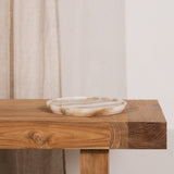 A rustic wooden console table with a thick rectangular top and single visible leg, styled with a small round stone tray. A beige curtain hangs to the left against a plain off-white wall.