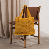 A mustard yellow crocheted tote bag with long shoulder straps, draped over a wooden chair with a brown leather seat in a minimalist room with beige curtains and a concrete floor.